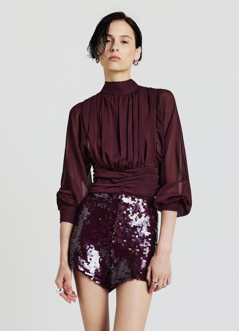 Blusa color bordeaux con dettagli plissettati, collo alto e maniche trasparenti abbinata a pantaloncini scintillanti con paillettes nella stessa tonalità.