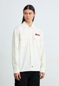 Cream button-up shirt with two front pockets, "Dickies" logo in red embroidery on the left chest, and a smooth fabric texture.