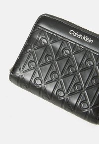Portefeuille en cuir noir avec un motif géométrique embossé, doté d'une fermeture éclair et de quincaillerie en argent. Logo "Calvin Klein" en haut.