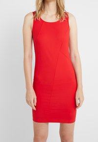 Robe moulante sans manches rouge en tissu extensible, avec un décolleté plongeant, une silhouette ajustée et des détails de coutures géométriques.