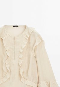 Blouse beige à manches longues avec fermeture éclair à l'avant et bordure à volants le long de l'encolure, de la poitrine et des poignets, fabriquée en tissu léger.