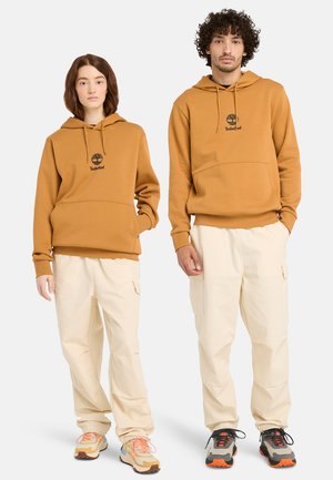Two models wear matching hoodies in mustard yellow with black logo, paired with cream cargo pants and sneakers in various colors.