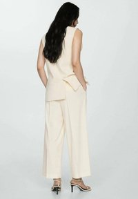 Top bej fără mâneci cu un design în straturi la spate, asortat cu pantaloni largi. Prezintă bretele subțiri pe tocuri negre. Textura materialului este netedă.