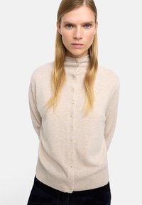 Beżowy sweter dzianinowy z wysokim kołnierzem i zapięciem na guziki z przodu. Miękka faktura, opadające ramiona i szczegół z ribbingu u dołu. Brak widocznych wzorów lub akcentów.