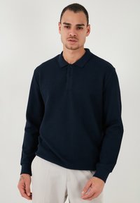 Polo cu mânecă lungă de culoare bleumarin, realizat din material cu caneluri, având gât cu nasturi și manșete, prezentat pe o persoană pe un fundal simplu.