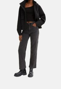 Giacca nera corta con cappuccio, indossata sopra un top nero aderente; abbinata a jeans larghi grigio scuro e stivaletti neri chunky.
