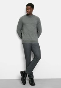 Grauer Pullover mit rundem Ausschnitt, kombiniert mit dunkelgrauen Hosen. Das Model trägt schwarze Schuhe. Glatte Textur, taillierte Passform und minimalistisches Design.
