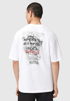 Personne vue de dos portant un t-shirt blanc avec un texte noir et rouge ainsi qu'un graphique de fil barbelé dans le dos.