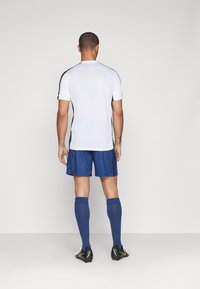 Wit sportshirt met zwarte zijaccenten, gecombineerd met blauwe shorts. Blauwe kniekousen en zwarte voetbalschoenen met oranje details maken de look compleet.