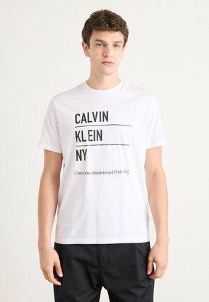 Fehér pamut póló fekete felirattal: "CALVIN KLEIN NY" és "Calvin Klein 1968-ban alapítva NYC" aláhúzva. Rövid ujjú és kerek nyakú.