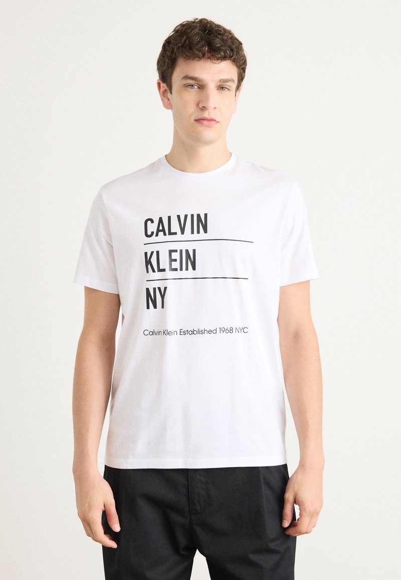 Bijela pamučna majica s crnim natpisom: "CALVIN KLEIN NY" i "Calvin Klein Established 1968 NYC" koji je podvučen. Kratki rukavi i okrugli izrez.