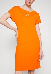 Kvinna i en klar orange kortärmad klänning med EA7 Emporio Armani-logga över bröstet, poserar med ena handen på höften.