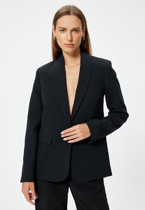 Blazer nero con rever a scialle, chiusura con un bottone, due tasche frontali e texture liscia, indossato sopra pantaloni abbinati.