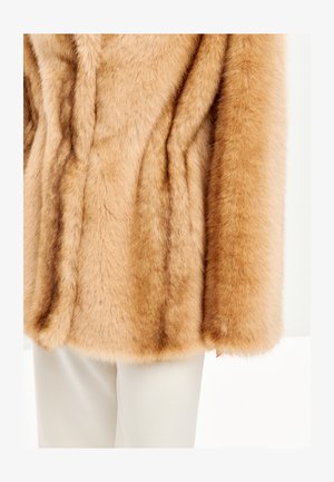 Abrigo de piel sintética en tono beige con una textura suave y esponjosa. Presenta mangas anchas y un acabado liso, combinado con pantalones blancos en el fondo.