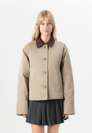 Veste cropped beige avec col marron, quatre boutons et poches latérales. Présente une texture lisse et une coupe décontractée avec des manches larges.