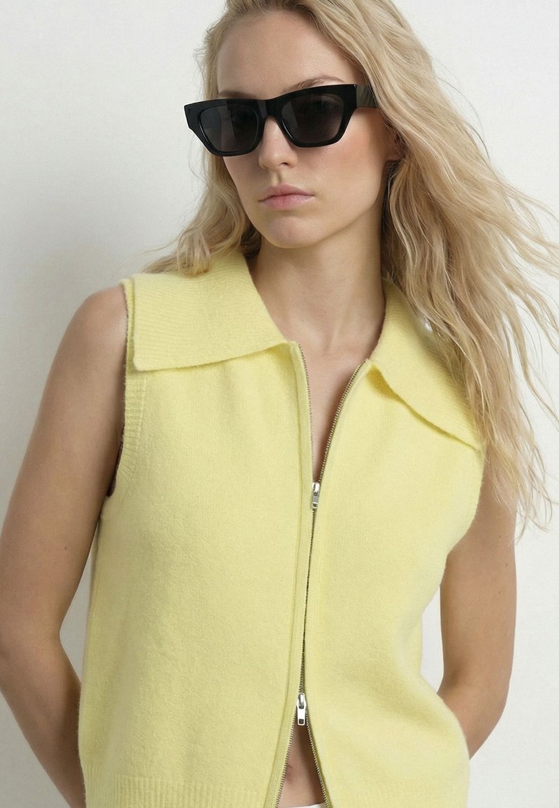 Femme blonde portant des lunettes de soleil rectangulaires noires et un haut zippé sans manches jaune pâle à large col, regardant légèrement sur le côté.