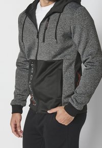 Hoodie zippé gris et noir avec une partie supérieure texturée, une partie inférieure lisse, des poches avant et une capuche avec cordon de serrage.