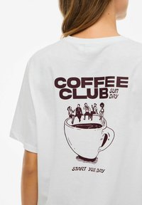 T-shirt bianco in cotone con un design stampato di una tazza di caffè con figure e il testo "COFFEE CLUB SUN DAY" e "INIZIA LA TUA GIORNATA."