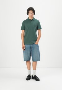 Polo vert à manches courtes, porté avec un short en denim arrivant aux genoux ; des mocassins noirs et des chaussettes grises complètent la tenue.