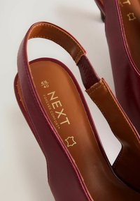 Bordó bőr pántos cipők barna párnázott talpbéléssel, melyen dombornyomott "NEXT FOREVER COMFORT" felirat látható. Közepes sarokmagasságú dizájn.