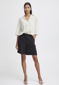 Chemise beige clair à boutonnières avec un col, deux poches avant et une coupe décontractée, assortie à un short cargo noir avec poches latérales.