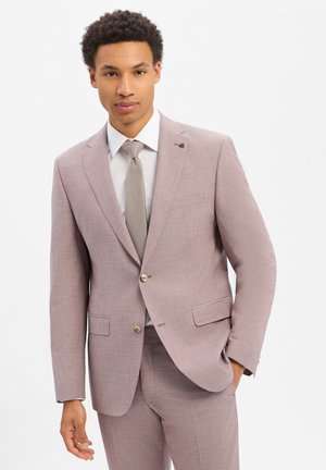 Junger Mann in einem hellen mauvefarbenen Anzug mit weißem Hemd und grauer Krawatte, der mit einer Hand in der Tasche vor einem schlichten weißen Hintergrund steht.