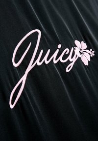 Melna auduma materiāls ar rozā izšūtu tekstu "Juicy", blakus burtiem ir florāla apdare.