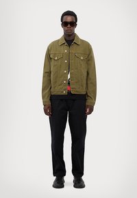 Olive green denim jacket with chest pockets, silver buttons, worn over a black shirt and black pants, paired with black sunglasses.