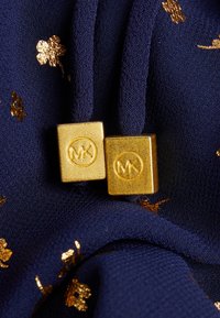 Guld MK-logohardware på en marinblå tyg med spridda guldblommönster, som visar en mjuk textur och en rik färgkontrast.