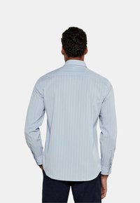 Camicia azzurra chiara a maniche lunghe con strisce bianche verticali. Presenta un colletto e polsini con bottoni, realizzata in un tessuto morbido. L'orlo è leggermente curvato.