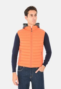 Uomo che indossa un gilet senza maniche arancione imbottito sopra una camicia a maniche lunghe blu navy e jeans blu, in piedi con una mano in tasca, mentre guarda a destra.