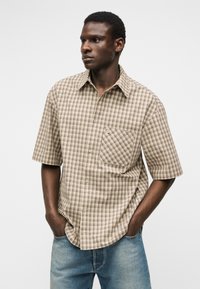Hombre con camisa beige de manga corta de cuadros, con bolsillo delantero, con las manos en los bolsillos, frente a un fondo liso y claro.