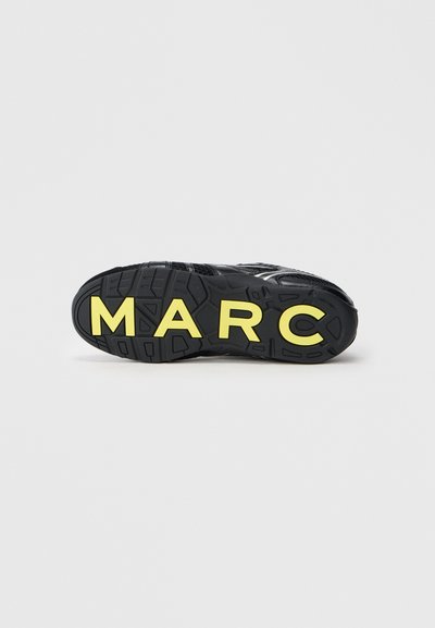 Suela de zapatilla negra con letras amarillas que deletrean "MARC" incrustadas en el diseño de la suela, mostrada sobre un fondo blanco liso.