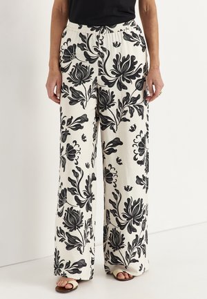 Løse hvide bukser med stort sort blomsterprint, båret med sandaler og en sort top, vist fra taljen til fødderne.