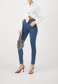 Camisa blanca de manga larga con nudo en el frente, combinada con jeans skinny azules. Tacones negros de tiras y un bolso pequeño texturizado completan el atuendo.