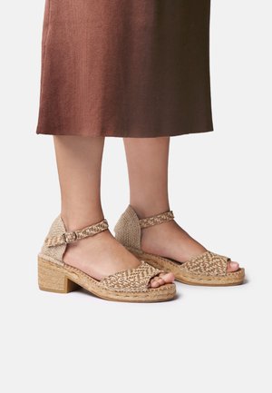 Sandali beige intrecciati con cinturino alla caviglia e tacco largo indossati con una gonna midi marrone su sfondo chiaro.