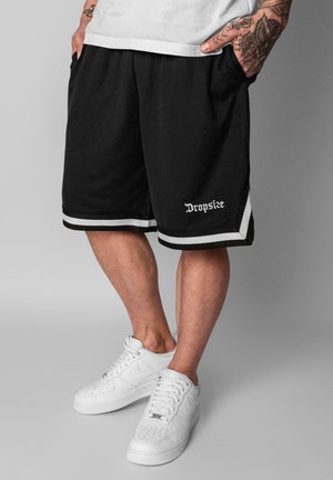 Man draagt zwarte basketbalshorts met witte rand en witte sneakers, handen in zakken, zichtbare tatoeages op onderarmen.