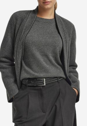 Frau trägt einen dunkelgrauen Strickpullover, eine passende offene Strickjacke, einen schwarzen geflochtenen Gürtel und hochgeschnittene schwarze Hose mit Händen in den Taschen.