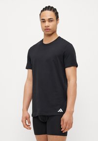 Fekete pamut póló rövid ujjakkal, kerek nyakkal, laza fazonban, az alsó bal oldalon fehér Adidas logóval. Az anyag simának tűnik.
