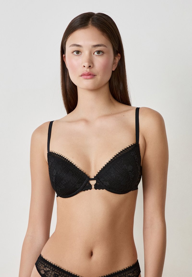 Reggiseno in pizzo nero con coppe imbottite, bordi ondulati e una decorazione centrale a forma di foro. Spalline sottili e regolabili con dettagli a motivo intricato.