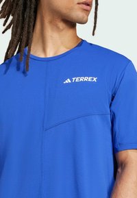 Blå idrottströja med korta ärmar, med en strukturerad paneldesign och vit "TERREX"-logotyp på bröstområdet.