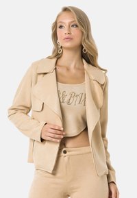 Veste en suède synthétique beige avec de grands revers, silhouette ajustée et coutures ton sur ton. Assortie avec un ensemble haut et bas coordonné.