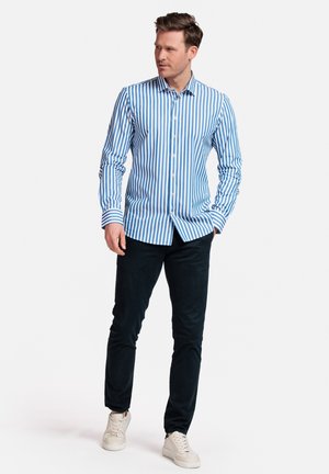 Chemise à rayures bleues et blanches à manches longues, associée à un pantalon foncé et des baskets claires. La chemise est dotée d'un col classique.