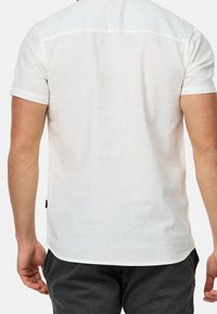 Mężczyzna w białej lnianej koszuli z krótkim rękawem i ciemnoszarych spodniach, stojący z rękami luźno wzdłuż ciała, plecami w stronę widza.