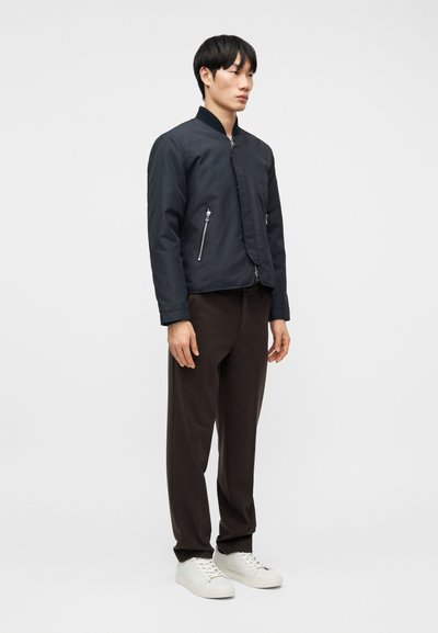 Άνδρας που φοράει μαύρο μπουφάν τύπου bomber, σκούρο καφέ παντελόνι και λευκά αθλητικά παπούτσια, στέκεται μπροστά σε λευκό, απλό φόντο.