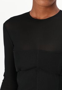Černý top s dlouhým rukávem vyrobený z lehkého materiálu. Obsahuje texturovanou část přes hrudník a vlnkovaný výstřih.