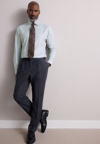 Světlounce zelená pruhovaná košile s hnědou vzorovanou kravatou, doplněná o na míru šité tmavě šedé kalhoty a černé boty. Stojící póza před neutrální zdí.