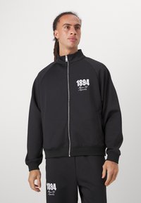 Svartid zip-up treningsjakke med høy krage og ribbestrikkede mansjetter, med hvit trykt tekst "1894" foran. Glatt stofftekstur.