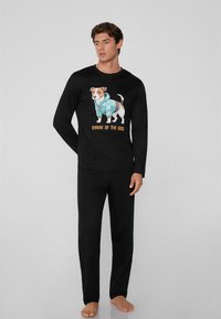 Schwarzes langärmliges Schlafanzugoberteil mit einem Grafiken eines Hundes in einem blauen Outfit und dem Text "VORSICHT VOR DEM HUND", kombiniert mit schwarzen Schlafanzughosen.