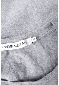 Šedé bavlněné tričko s kulatým výstřihem, na límci je bílá etiketa se nápisem "CALVIN KLEIN JEANS" a velikostí 'M'.